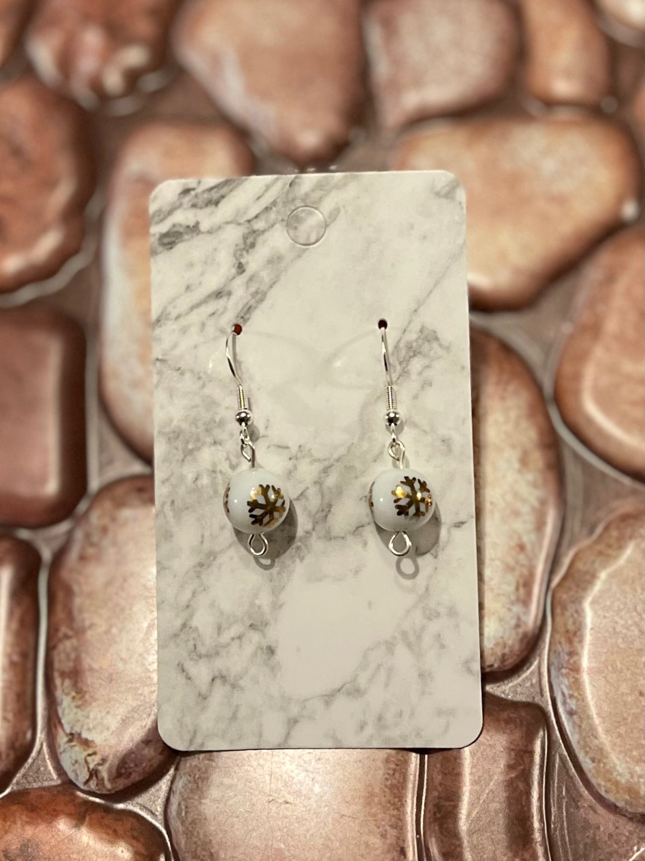 Pair of Sterling Silver Christmas earrings. Dangle/Drop style earrings with gold snowflakes on a white bead. Pictured on a marble earring card with a stone background.