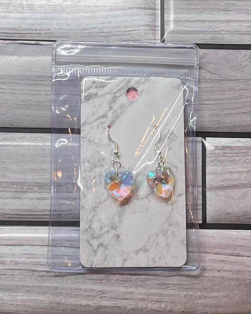 Heart-shaped earrings in clear packaging on a marble card with a gray tiled background